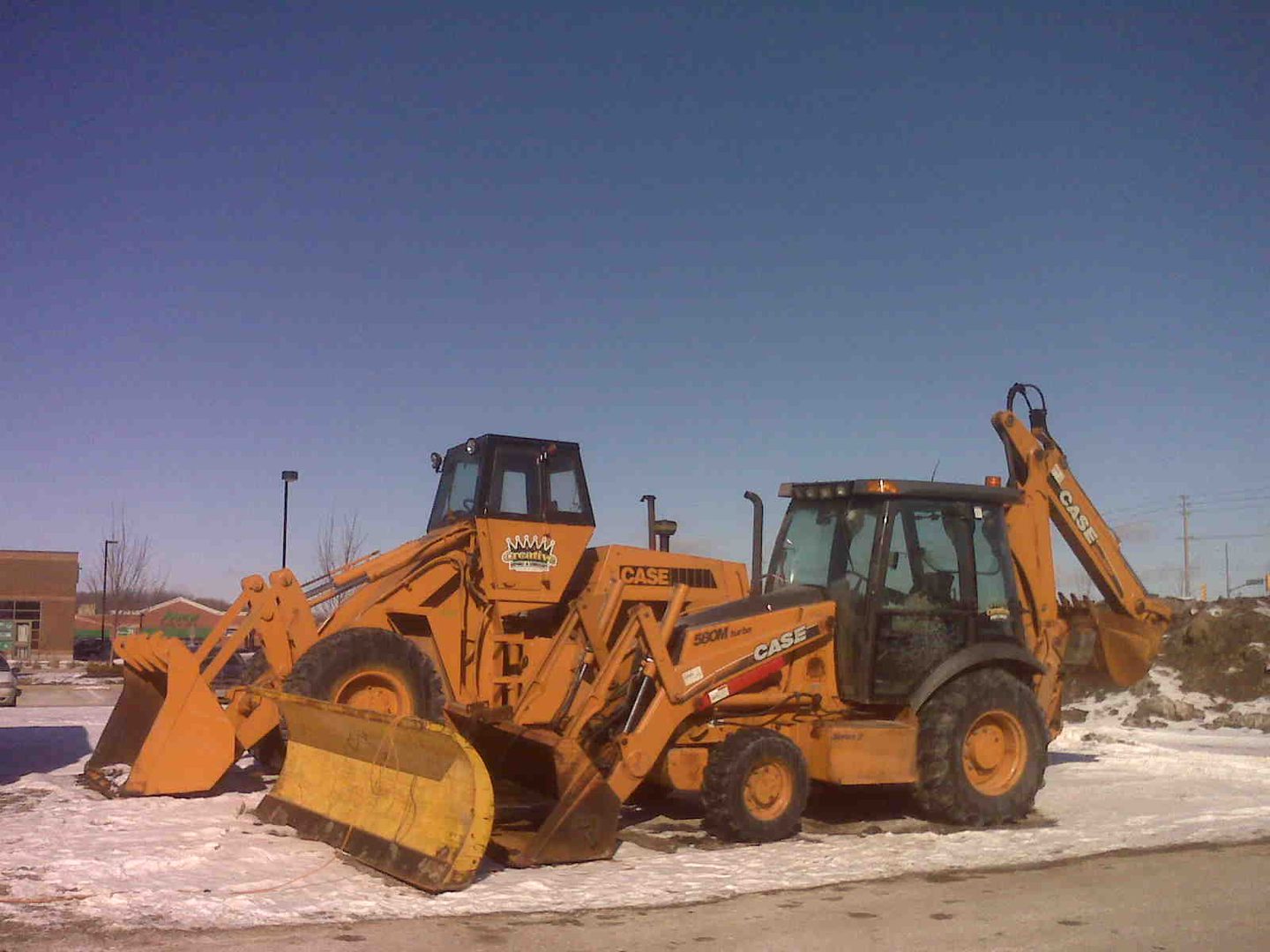 A yellow tractor parked in the snow near some other vehicles.