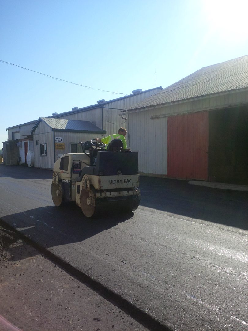 A man in yellow jacket riding on the back of a road roller.